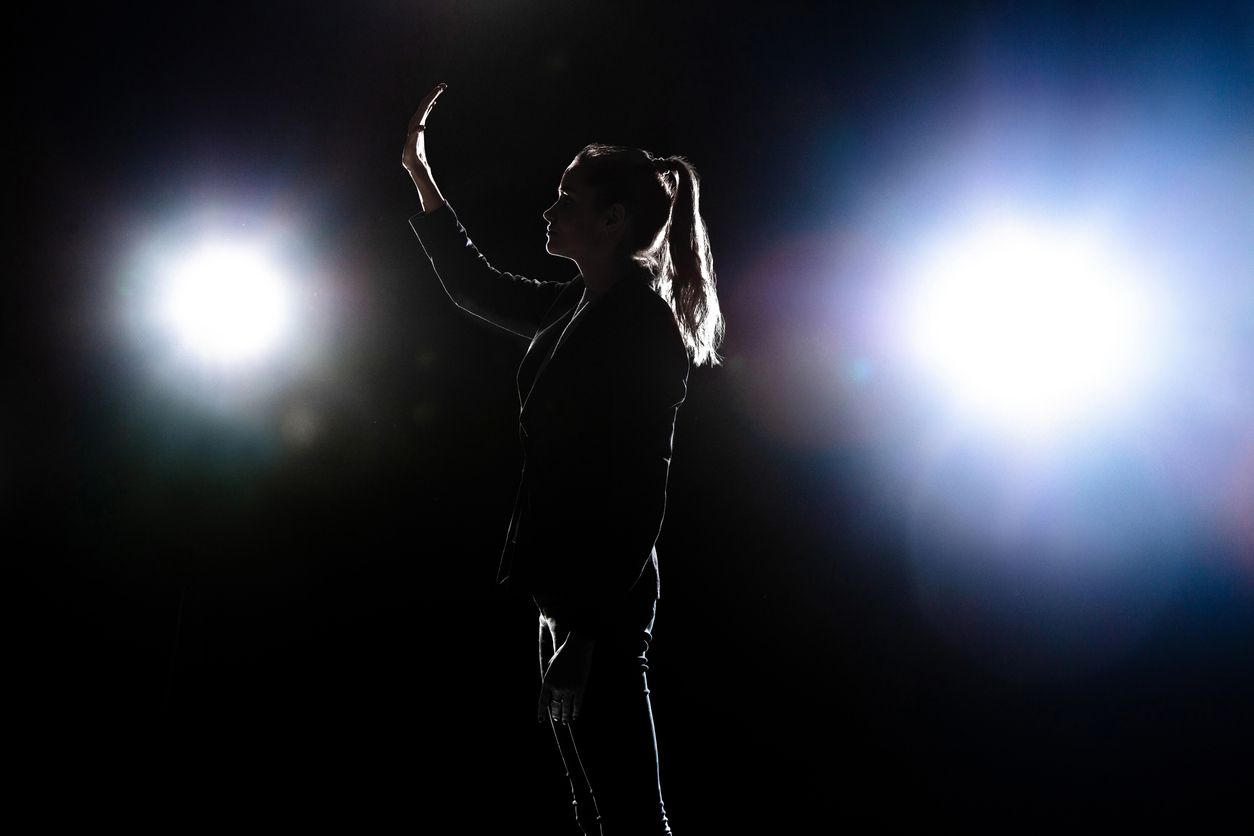 Silhouette d'une femme politicienne sur une scène, devant des projecteurs, avec une main levée.