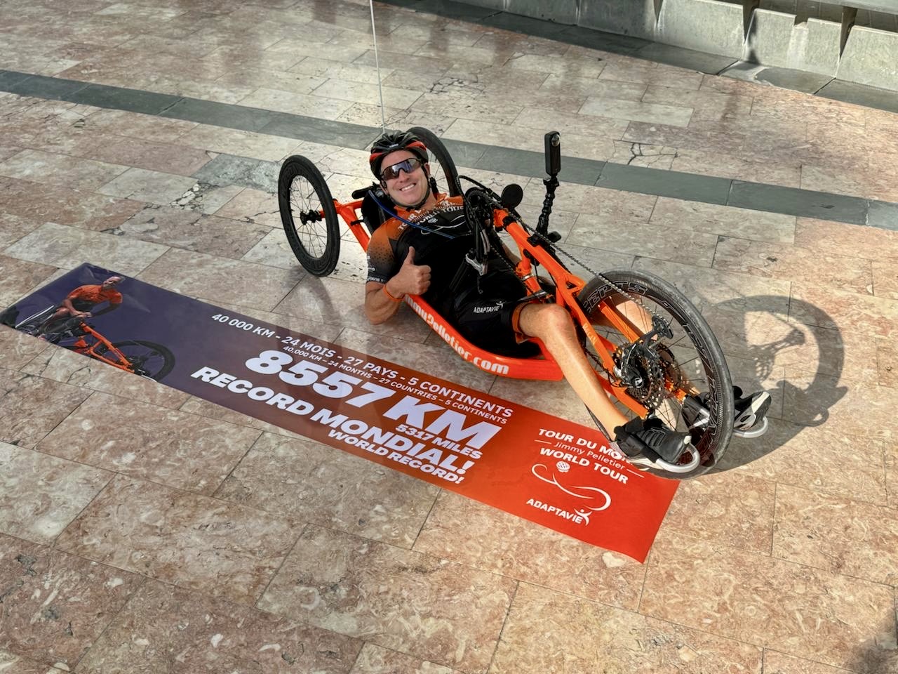 L'athlète paralympique Jimmy Pelletier installé sur son vélo à main devant une banderolle soulignant l'atteinte de son record mondial de 8557 km parcourus.